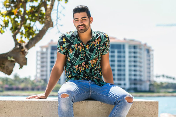 Ainda com cabelo preto, Dilsinho posa para foto ao lado de árvore. Cantor está sentado em uma mureta, com as mãos apoiadas