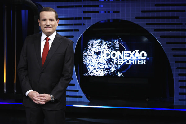 Logo do programa Conexão Repórter à direita, com Roberto Cabrini, vestindo terno completo, à esquerda