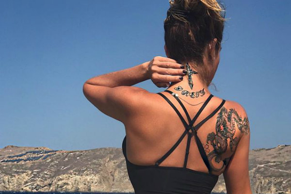 Com um maiô preto e de costas para foto, Luize Altenhofen levanta o cabelo para mostrar as tatuagens