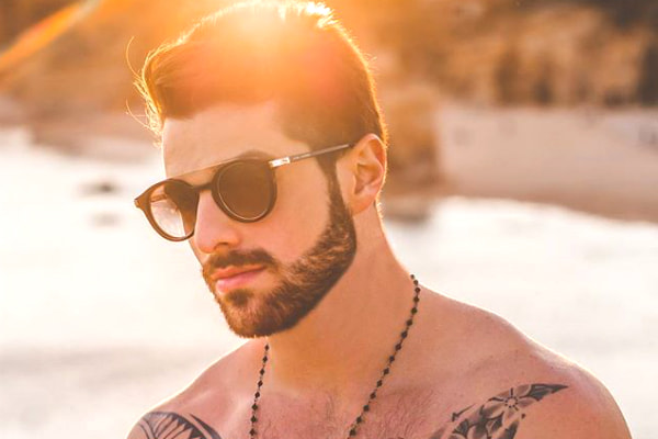 Com sol alaranjado ao fundo, DJ Aloz aparece sem camisa, de óculos escuros e com colar, na praia