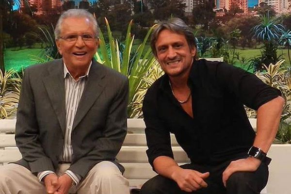 Carlos Alberto de Nóbrega e o filho Marcelo de Nóbrega sentados no banco do programa A Praça é Nossa sorrindo