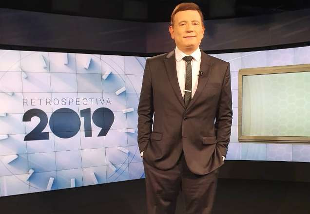 Roberto Cabrini posa para foto ao lado do logo da retrospectiva 2019 do SBT