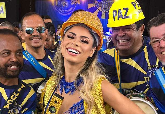 A cantora Lexa sorrindo. Ela está fantasiada de arquiteta, pois está em um ensaio de carnaval. Ela está sorrindo. Atrás, os músicas da escola de samba