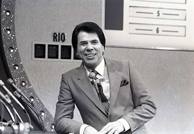 Na imagem preto e branco, o apresentador Silvio Santos está sorrindo apoiado em uma bancada. O registro é do ano de 1984