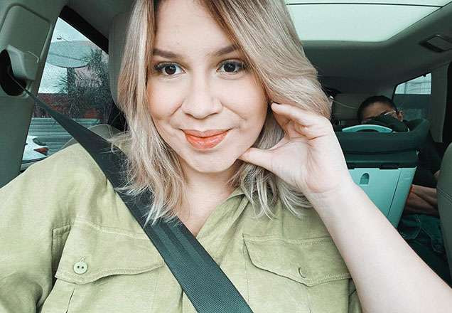 Marília Mendonça está com a mão no rosto sorrindo. Ela está usando uma blusa verde, e está sentada no banco de um carro