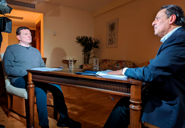 Em um quarto de hotel, Roberto Cabrini e Wassef - cada um de um lado da mesa - conversam e são filmados por uma câmera