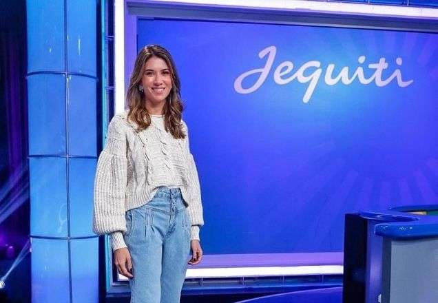 Rebeca está de pé, usando um tricô branco e uma calça jeans clara e posa para a câmera na frete de um painel da Jequiti | Foto: Gabriel Cardoso/SBT