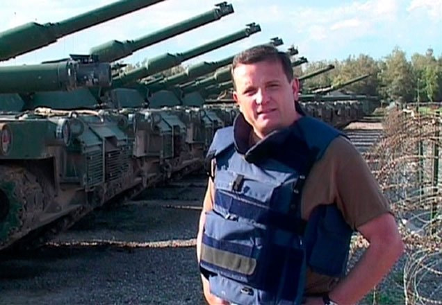 Com tanques de guerra ao fundo, Robert Cabrini, que usa colete à prova de balas, conta episódio marcante no Iraque