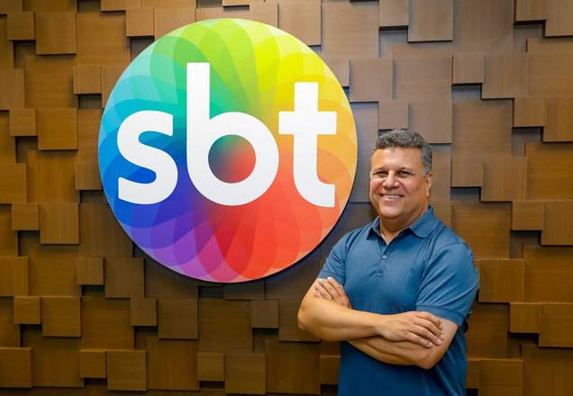 De braços cruzados, Téo José sorri para a foto com o logo do sbt ao fundo