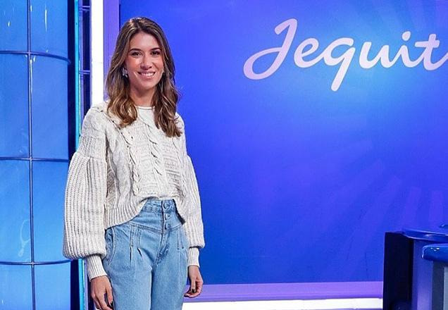 Rebeca está de pé ao lado de um telão com a palavra Jequiti, ela usa blusa bege e calça jeans | Foto: Gabriel Cardoso/SBT