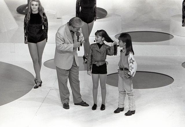 Foto em preto e branco mostra Gugu, Sandy e Junior no palco do Domingo Legal