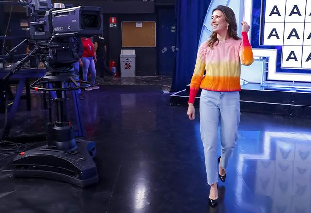 Rebeca anda pelo palco do Roda a Roda, atrás dela está o painel cheio de letras "a" e ao lado, uma câmera de televisão. Ela veste jeans e tricô colorido | Foto: Gabriel Cardoso/SBT