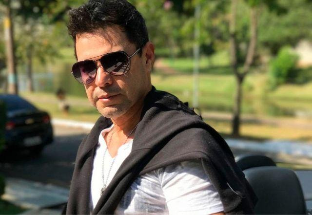 O cantor Zezé Di Camaro usa óculos escuros e blusa de frio amarrada ao pescoço. Ele sorri.