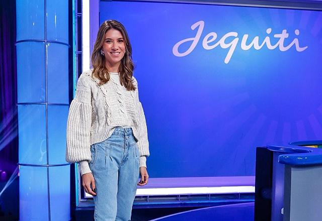 Rebeca usa tricô bege, calça jeans clara, salto alto laranja e posa sorrindo de pé para foto na frente de um telão escrito Jequiti Foto: Gabriel Cardoso/SBT