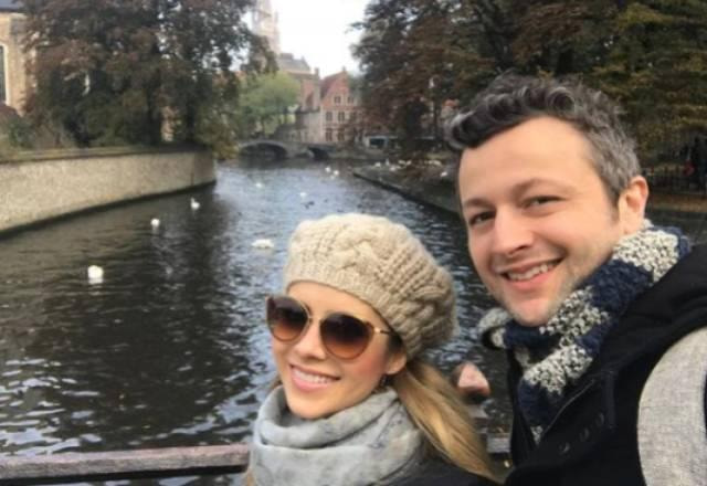 Sandy está sorrindo e vestida de boina de tricot bege, cachecol cinza e um óculos escuro. Ela está ao lado de Lucas Lima, que está de cachecol cinza e azul e moletom. Os dois estão na frente de um canal e aparece uma ponte ao fundo.