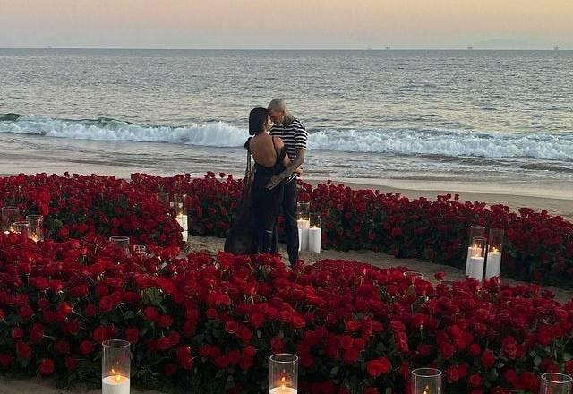 Kourtney de vestido preto, Travis de calça preta e camisa listrada. Os dois abraçados dentro de um coração gigante de flores com velas em uma praia.