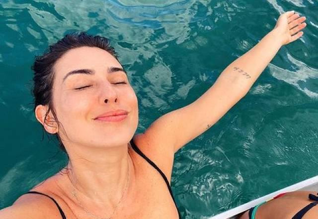 Fernanda Paes Leme de olhos fechados, com os braços abertos, com a cabeça olhando para o céu. De biquini preto, em cima da canoa, em um mar.