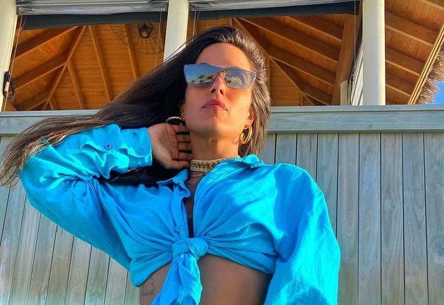 Carol Peixinho com blusa azul e óculos espelhados. Posando com a mão no pescoço e com o sol iluminando.