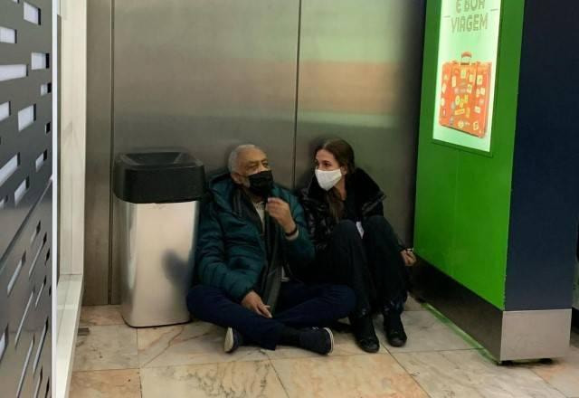 Gilberto Gil e Flora Gil esperam voo sentados no chão do aeroporto