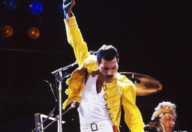 Vocalista Freddie Mercury durante show usando jaqueta amarela e com a mãe para cima
