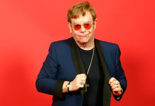 elton john posa em estúdio com fundo vermelho e terno azul