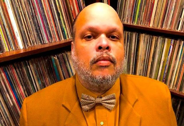 Ed Motta veste um terno amarelo, usa gravata borboleta e está em sua biblioteca
