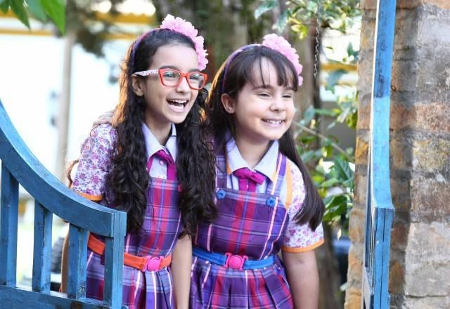 Frida e Bárbara vestidas com o uniforme do colégio Doce Horizonte, no portão da fazenda, rindo bastante e olhando para o lado