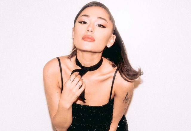 Ariana Grande veste um top preto colado ao corpo. Ela está em um estúdio fotográfico