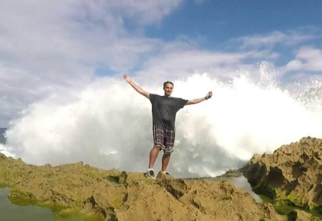 Homem em cima de umas rochas, abrindo os braços, posando para uma foto, com o mar atrás e uma onda batendo nas pedras