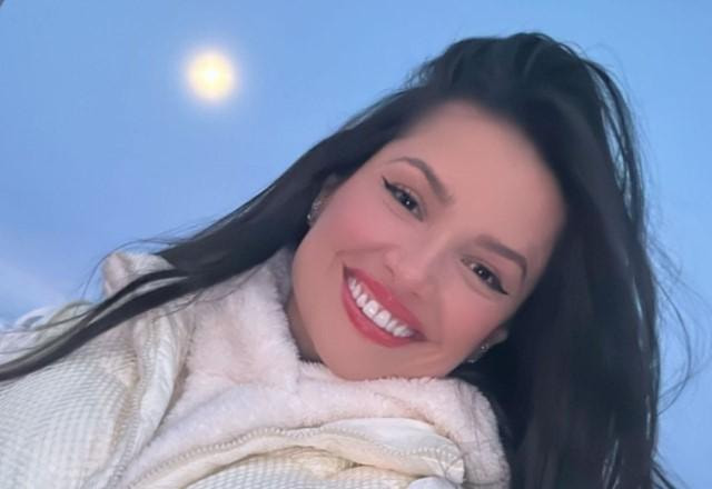 Juliette sorrindo, com roupa de inverno e a lua aparecendo no céu atrás