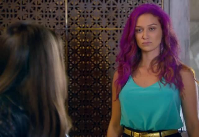 Regina com cabelo roxo e expressão de raiva em segundo plano, Isabela em primeiro plano, mas de costas