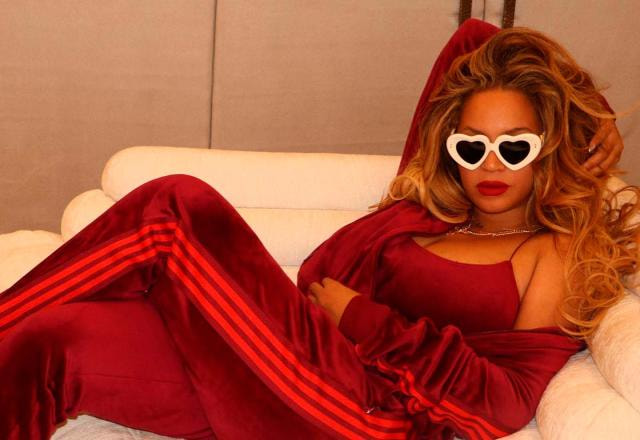 Beyonce deitada em um sofá de luxo, veste uma roupa vermelha, batom e óculos