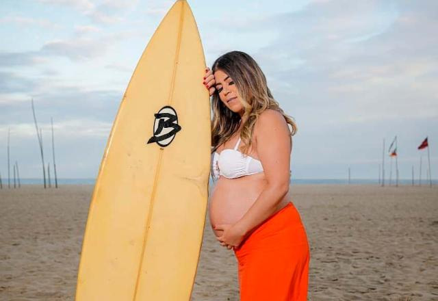 Raquel Pacheco, a Bruna Surfistinha, grávida, apoiada em uma prancha de surfe na praia