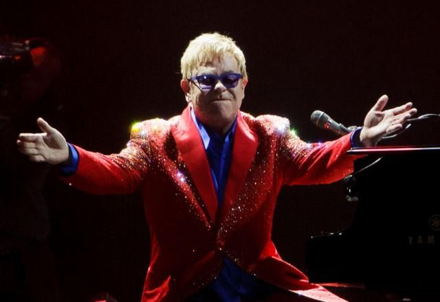 Elton John sentado em frente a um piano, no palco, e usando terno vermelho