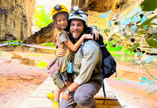 Com uma caverna ao fundo, a personagem Anna aparece sorrindo abraçada a seu pai, que também está sorrindo