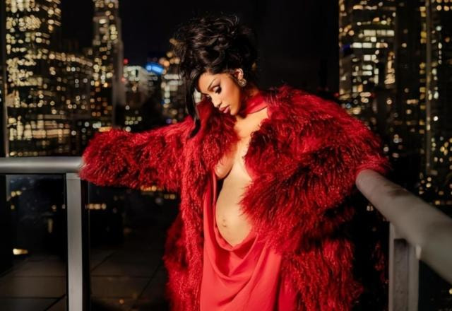 Cardi B vestida de vermelho gravida de seu terceiro filho olhando para sua própria barriga em uma sacada de prédio