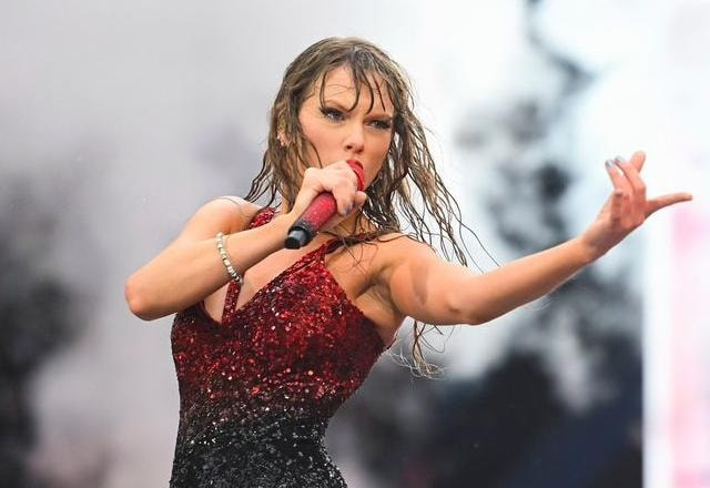 Foto de Taylor Swift em show, esta chovendo e a a cantora esta vestindo uma roupa vermelha, segurando o microfone e apontando para frente