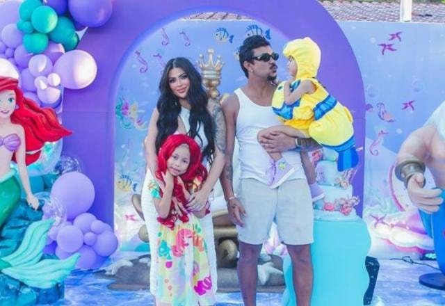 Xamã e Renata Gutierrez vestidos de branco, com Xamã segurando a filha Hanna no colo