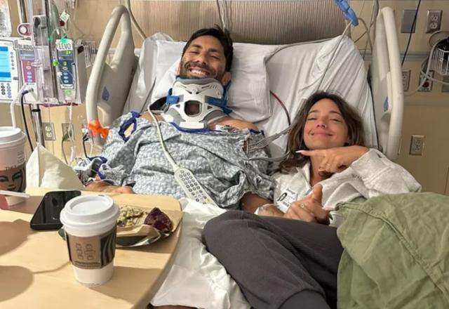 Nev Schulman em uma cama de hospital sorrindo para a foto, com sua esposa ao lado