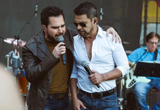 Zezé Di Camargo abraçado com Luciano em palco. Lucianovestido com uma jaqueta marrom e com o microfone em mão e Zezé Di Camargo com camiseta branca, ou vindo o irmão