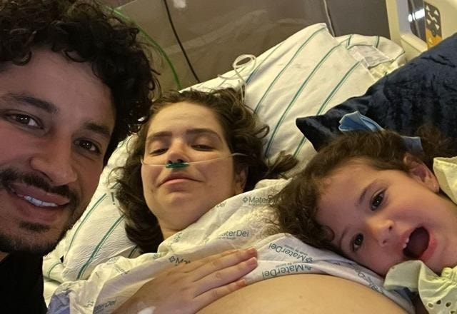 Foto de Camila Campos em cama hospitalar, grávida de sua filha Sofia, ao lado de seu esposo Léo e sua filha Bela