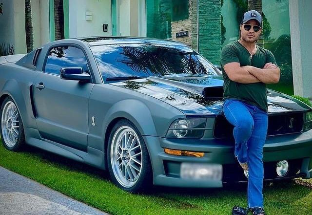 Eduardo costa vestido de boné, calça jeans, camisa verde e óculos escuros apoiado em seu carro, um mustang cinza