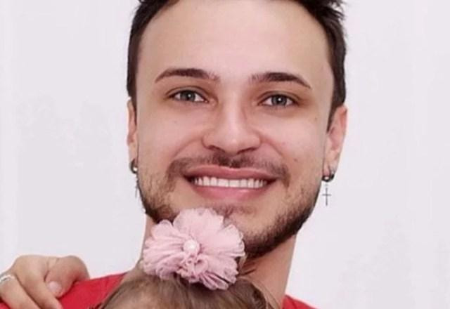 Imagem de Igor Viana sorrindo, vestido com uma camisa vermelha, segurando sua filha com paralisia cerebral