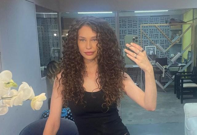 Imagem de Maidê Mahl, vestida de preto, com o cabelo cacheado e solto, segurando um celular em frente a um espelho