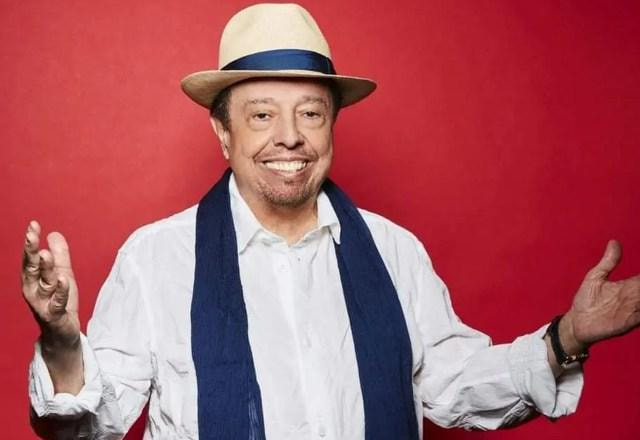 Sergio mendes vestido de branco, com um chapeu e o fundo em vermelho