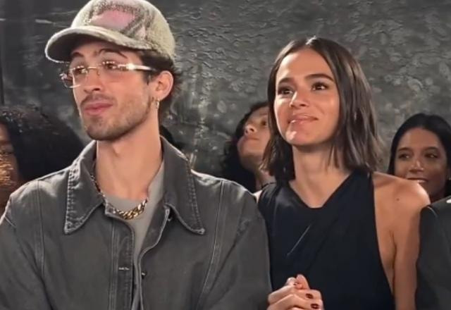 Imagem de Bruna Marquezine junto a João Guilherme, em um desfile de moda. Bruna vestida de preto e João Guilherme com boné e óculos