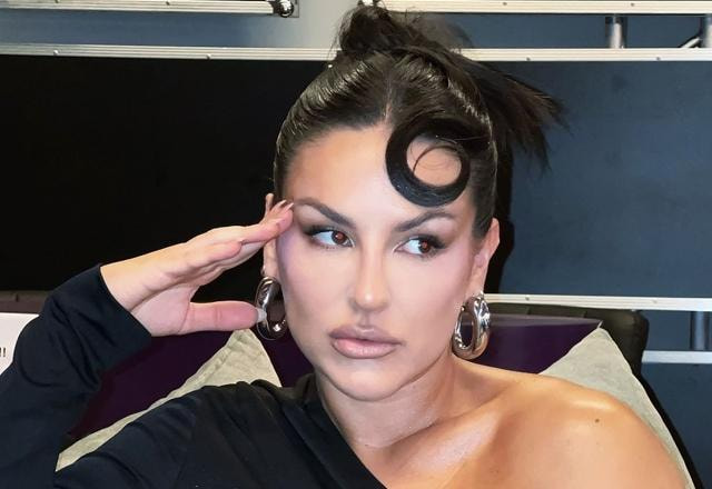 Imagem de Kefera, bem maquiada, com expressão facial marcante e olhando para a direita, com a mão no rosto