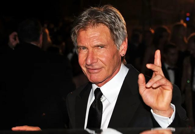 Foto de Harrison Ford vestido de terno, ao lado de uma carro, acenando com os dedos