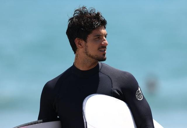 Imagem de Gabriel Medina, olhando para o lado, com o mar ao fundo, segurando uma prancha de surf, com o rosto sério