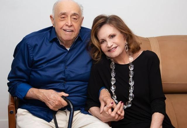 Foto de Rosamaria Murtinho junto com o esposo Mauro Mendonça de mãos dadas e sentados em um sofá marrom
